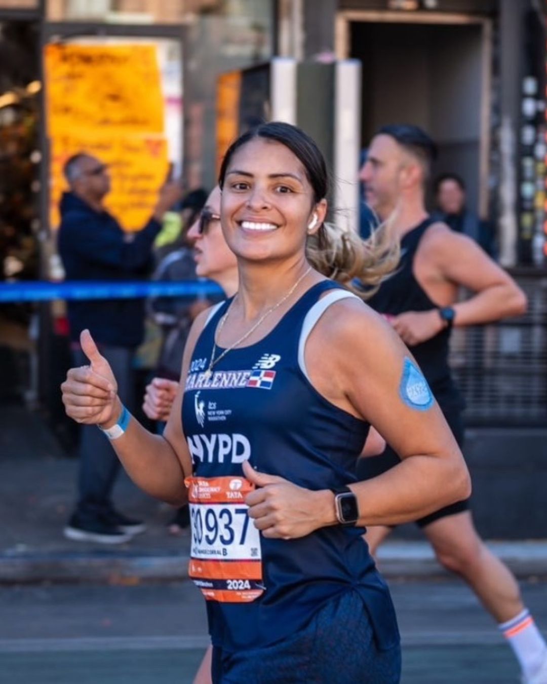 Woman running a marathon, giving a thumbs-up, with 'NYPD' and 'New Balance' branding.